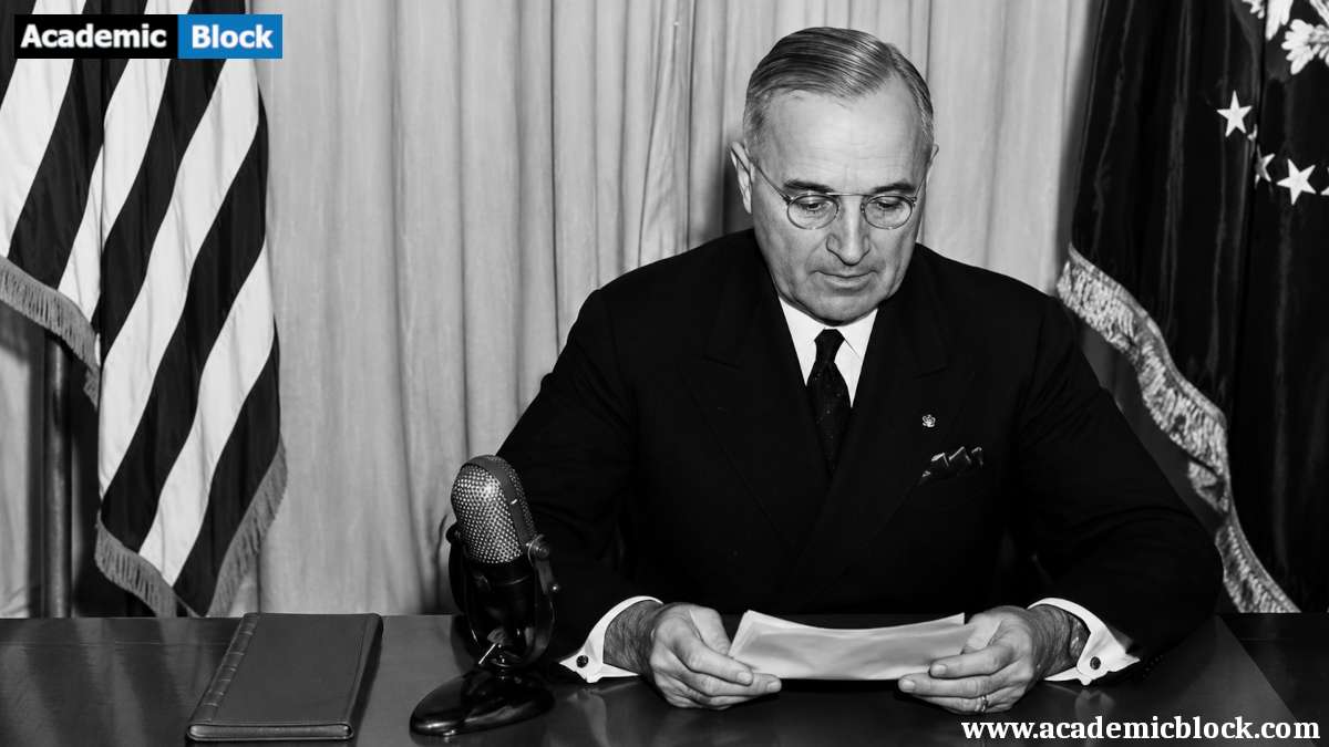 President Harry S. Truman seated at a desk reading documents and flags in the background.