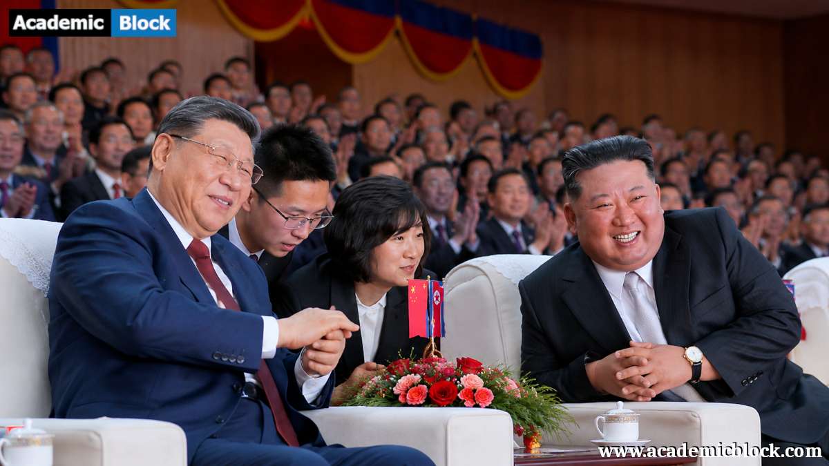 Xi Jinping and Kim Jong Un in formal suits smile and converse while seated at a public event, with a large applauding audience in the background.
